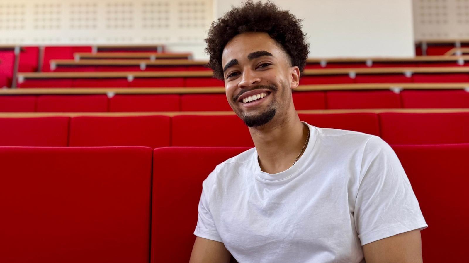 Kieran McRae is in a white T-shirt seated in a lecture theatre with rows of bright red tiered seats. The image is framed as a portrait, with the seating stretching into the background of the auditorium.