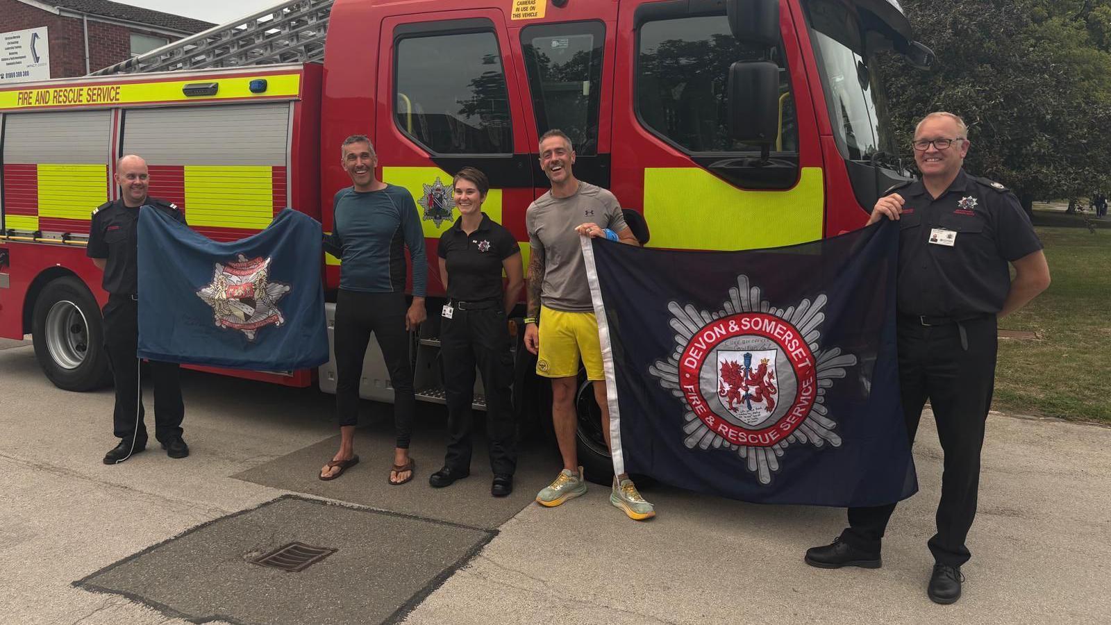 Three firefighters standing alongside two men in sportswear, who are holding fire service banners. They are standing in front of a fire engine.
