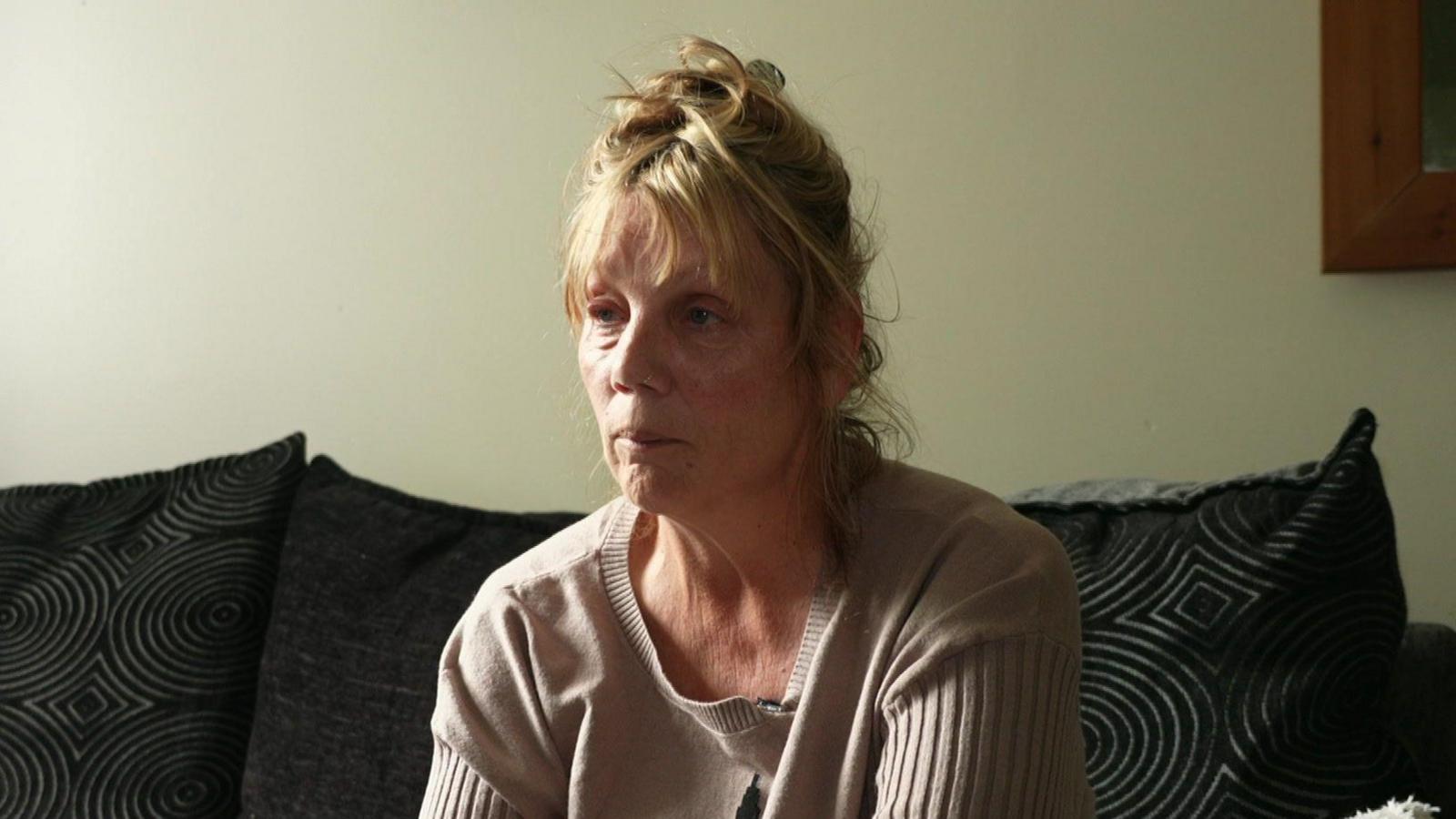 A woman sat on her sofa looking away from the camera rather upset. She has blonde hair and a beige sweater on. Her sofa has a few black cushions. It's pushed up against a clear white wall with a mirror to the right hand side.