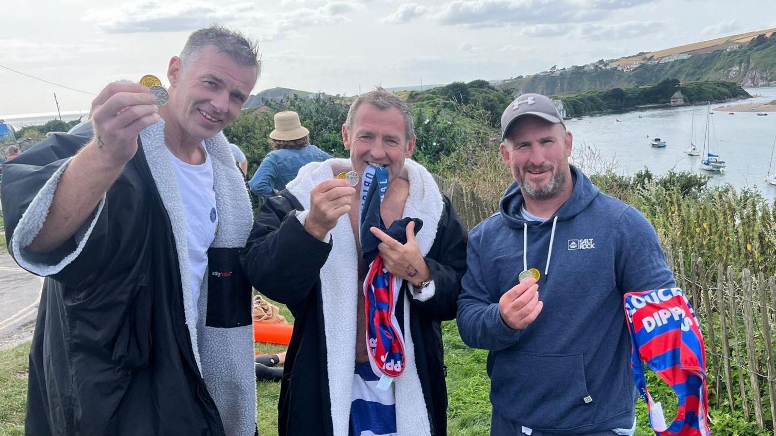 Three men standing by a lake wearing dry robes and holding speedos. They are posing with medals.