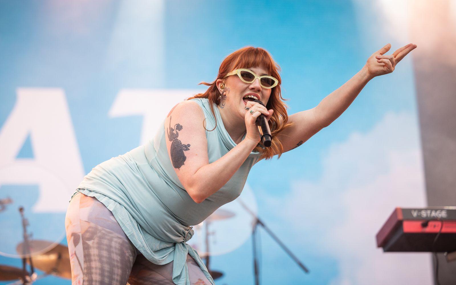 CMAT performing on stage wearing a baby blue dress, patterned tights and white sunglasses with her arm outstretched