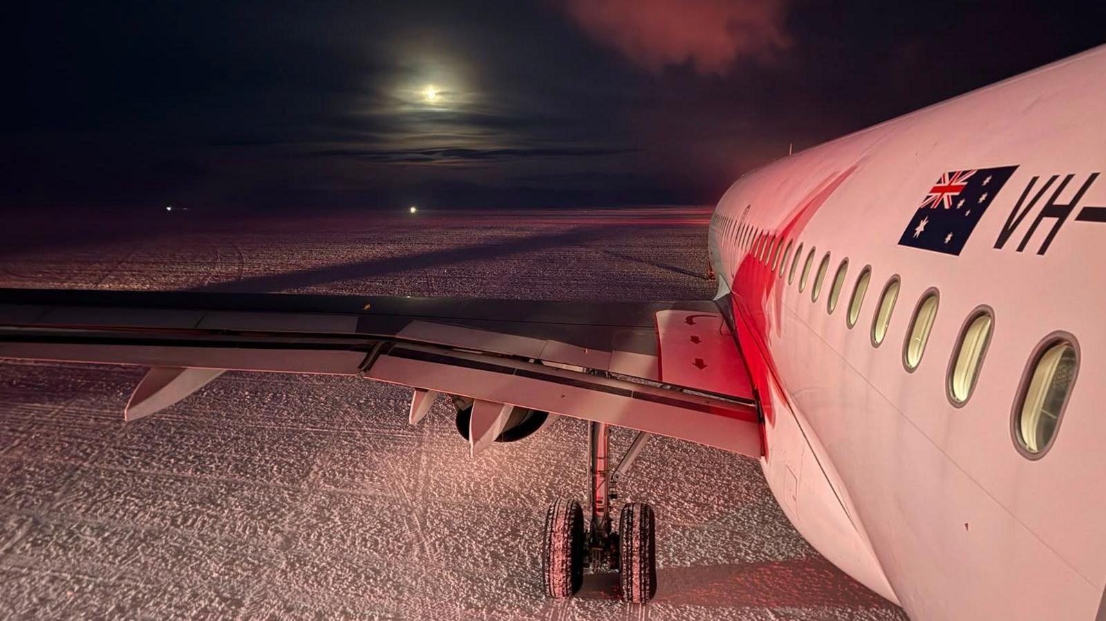 An aeroplane sits on the snow in the dark with the moon seen through the clouds