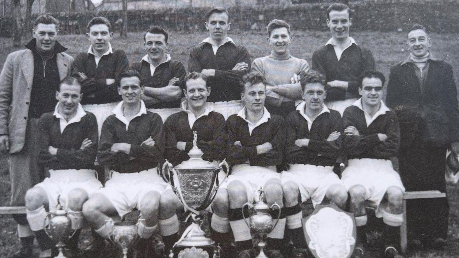 A group of footballers, in a black and white photograph, line up behind trophies