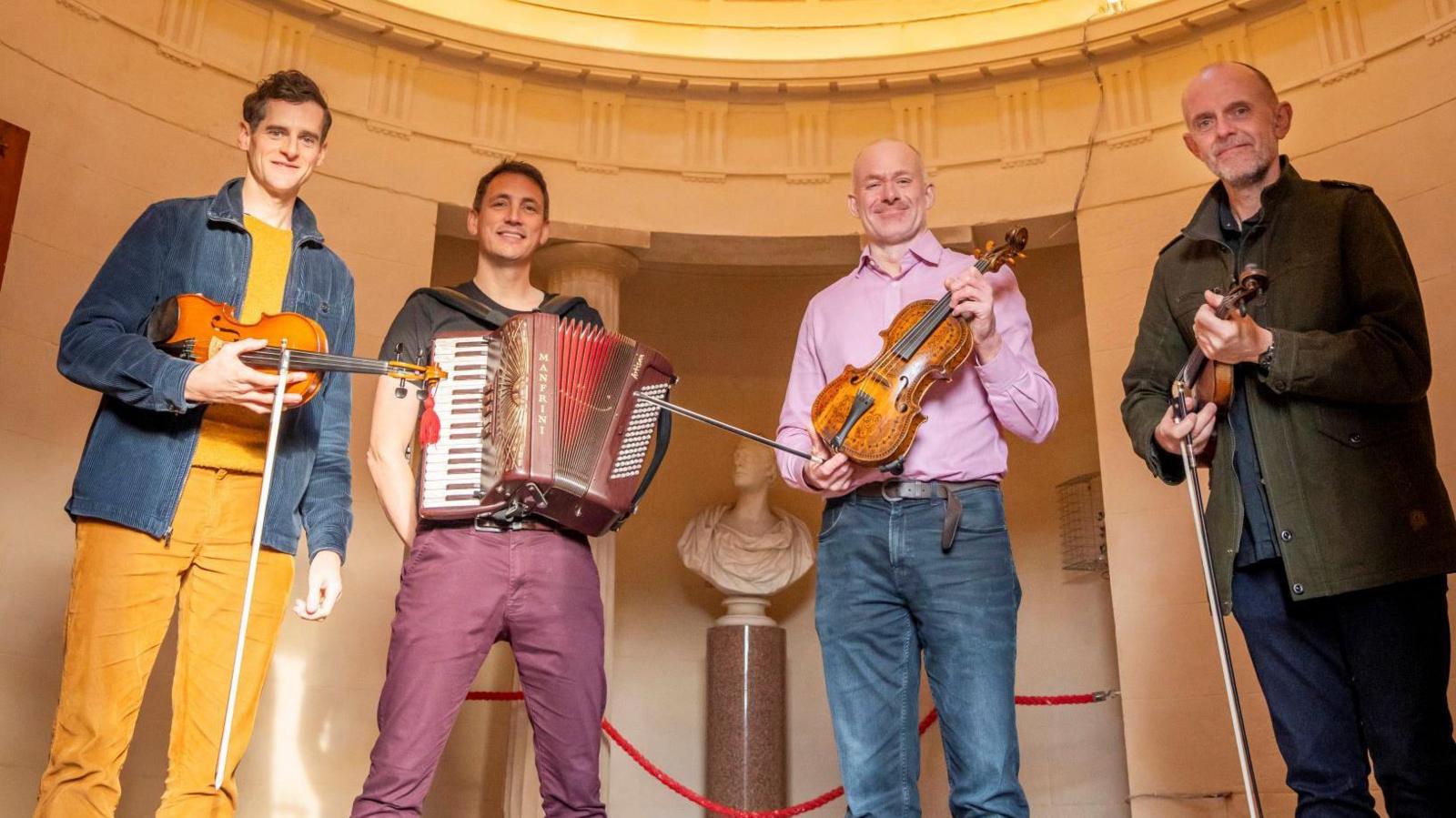 Ewen Henderson, Gary Innes, Alistair McCulloch and Duncan Chisholm standing inside the Burns Initiative, with a white marble bust of Burns behind them. They are all holding fiddles or accordions while looking towards the camera.