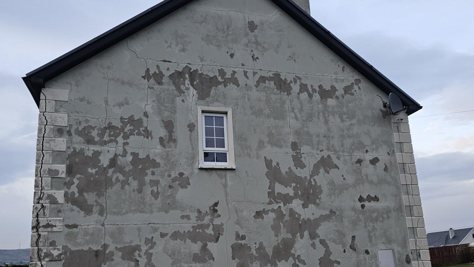A side profile of a grey house with a number of visible cracks and marks in the wall. 