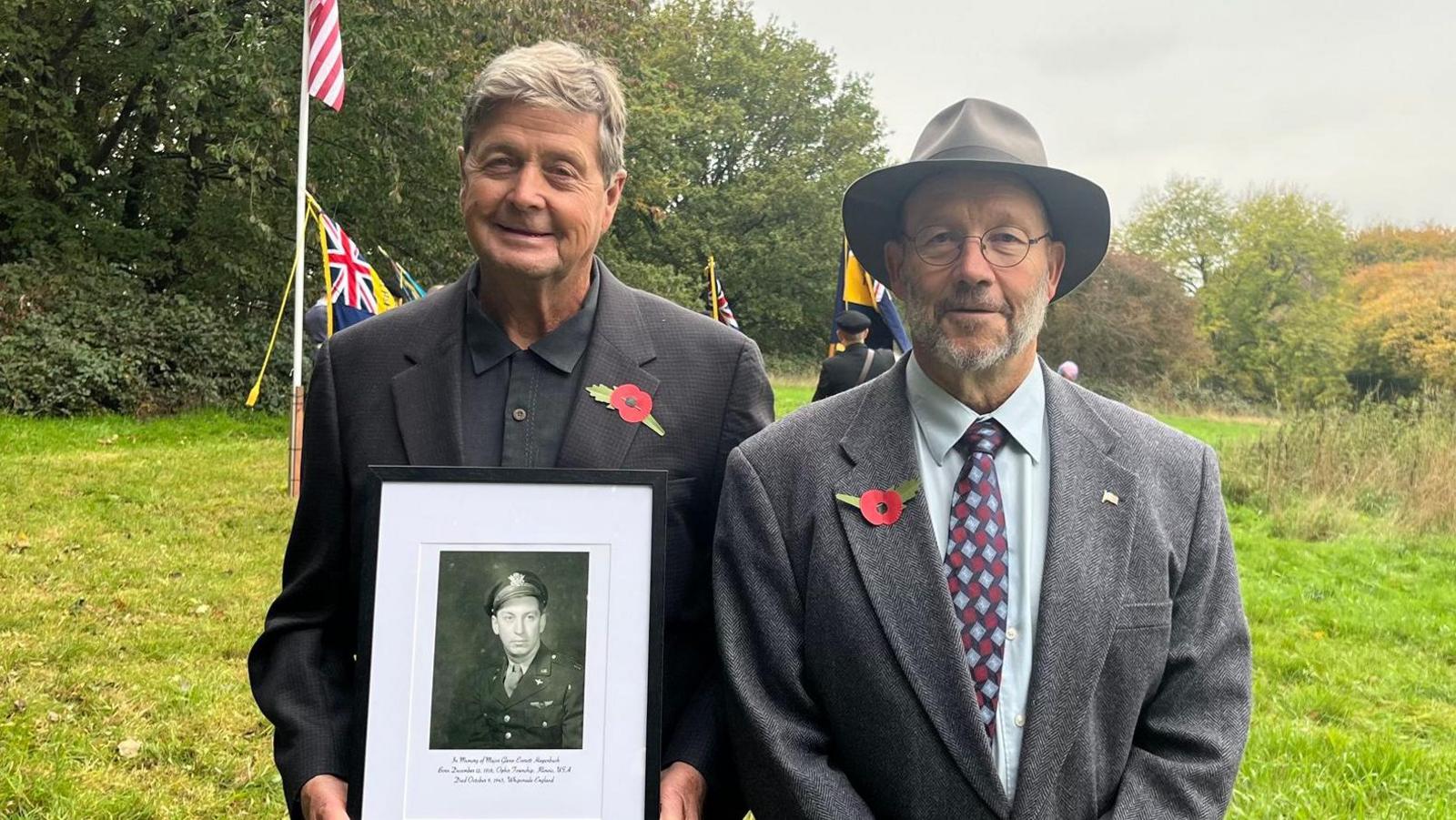 Two men stand in a field holding a framed photo of Major Glenn, in uniform. There is a flagpole behind them with a US flag. They both wear dark suits and poppies on their lapels. It is autumn.