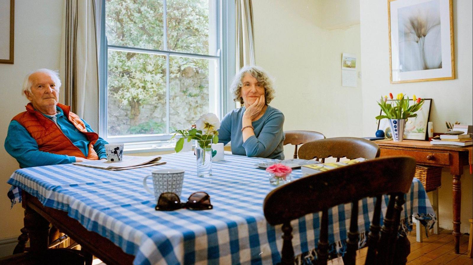 A man and a woman sit at a table covered in a blue gingham cloth. There are three mugs and a vase of flower sat on top and they are both smiling at the camera. In the background is a large window through which trees are visible