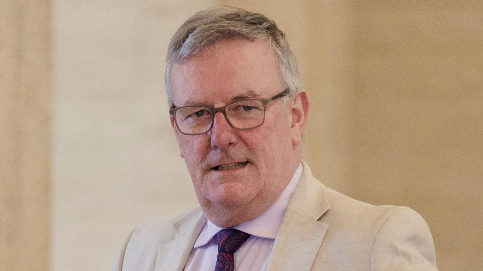 Nesbitt with short grey hair, square metal glasses looks to his left with his mouth slightly open showing his teeth. He is wearing a light cream suit, pink shirt and purple and pink patterned tie. The background, which is a dark cream wall, is blurred.