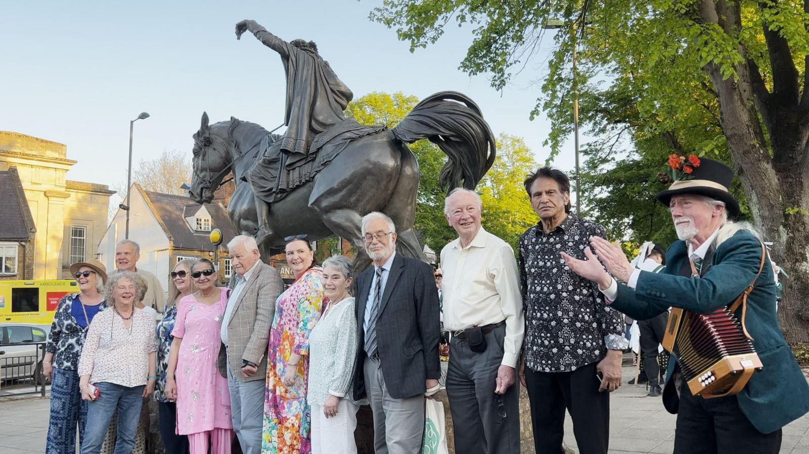 Project committee member and musician Donald McCombie gathered around the statue. Mr McCombie is clapping. It's a sunny day.