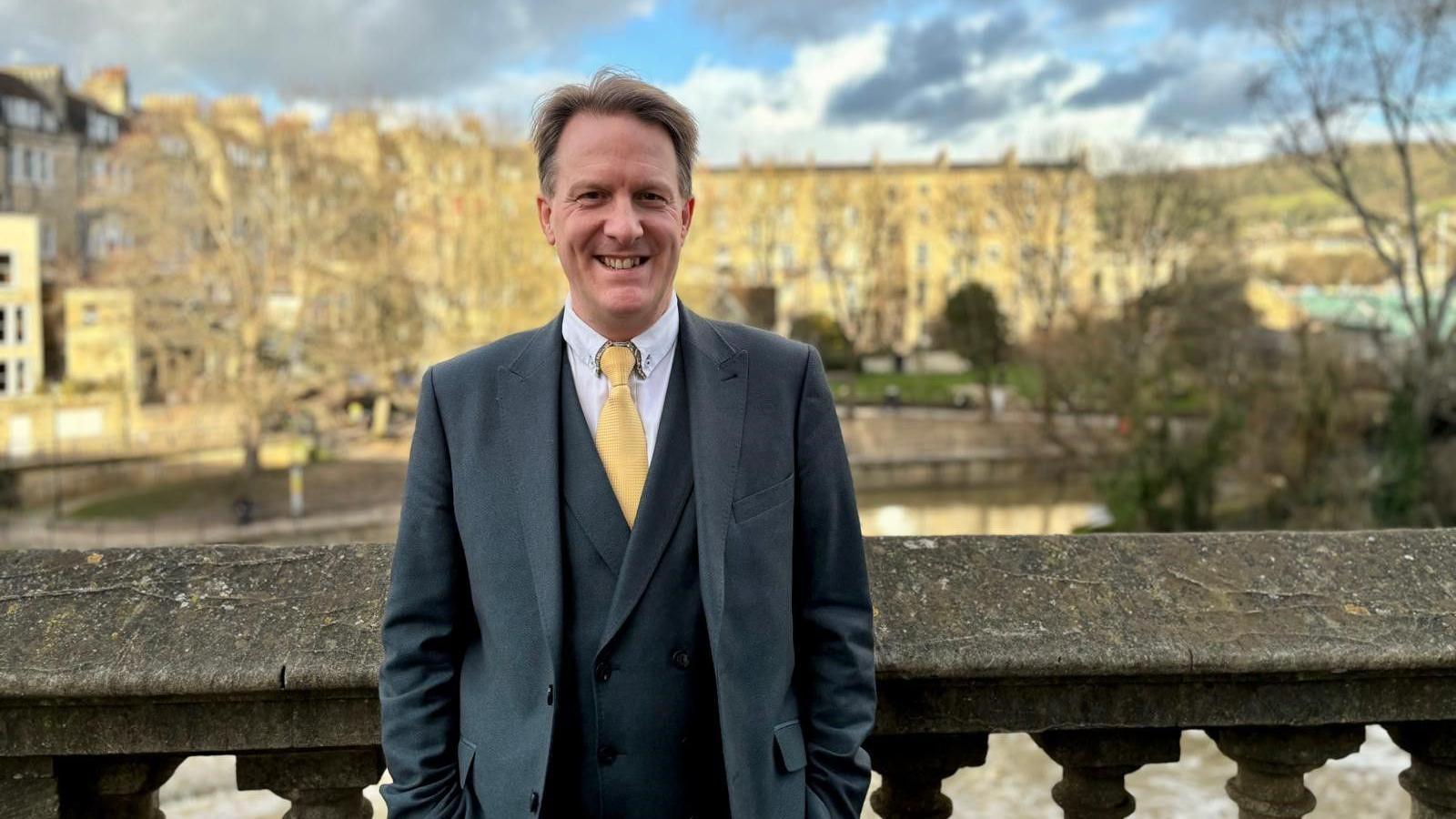 John Elliott, a middle aged man with brown hair, wearing a black suit and long black coat with a white shirt and yellow tie, smiles as he stands above the River Avon in Bath