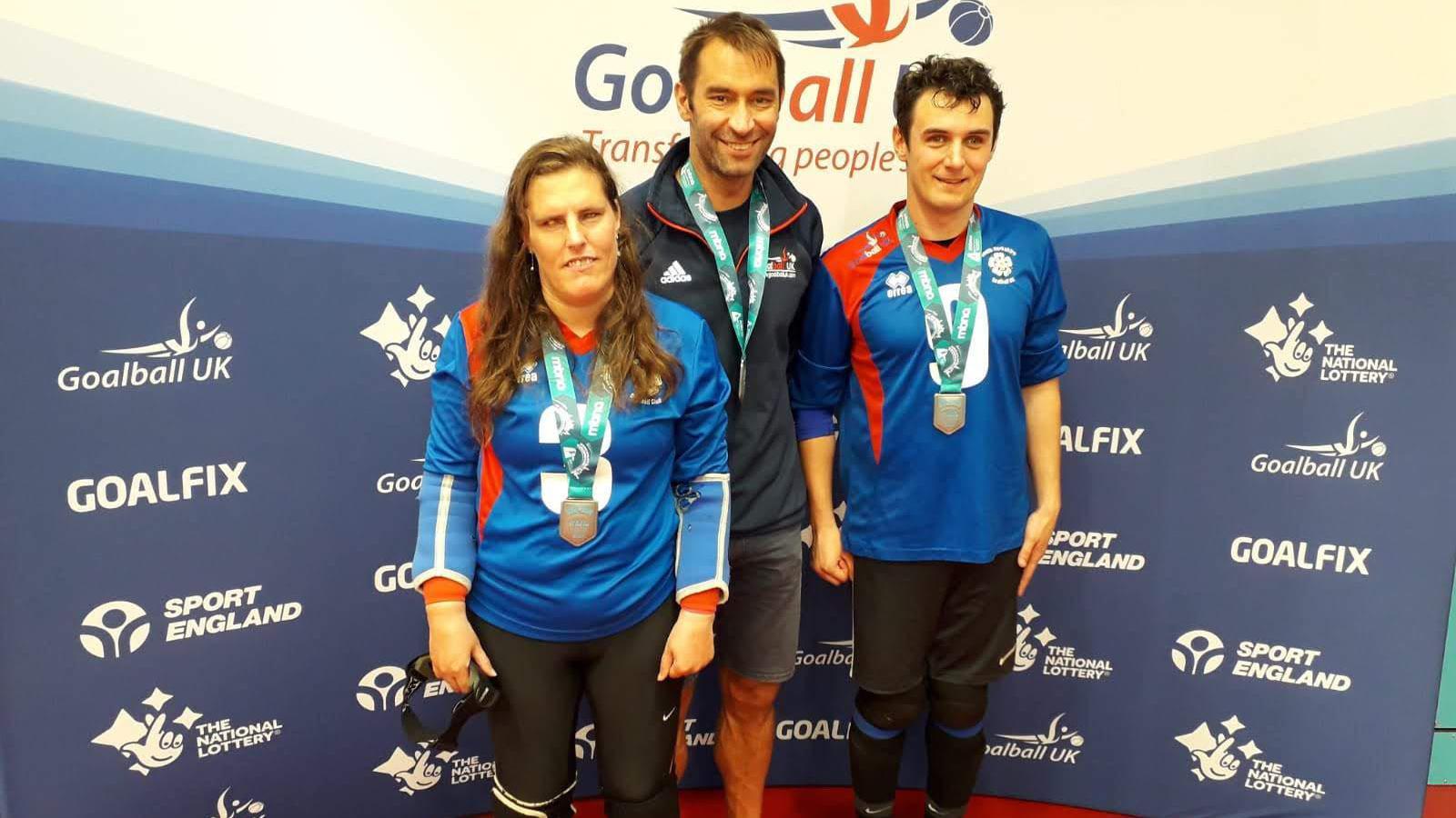 Two men and a woman pose for the camera wearing silver medals around their necks. The woman and one of the men are wearing a blue sports top and black lycra trousers