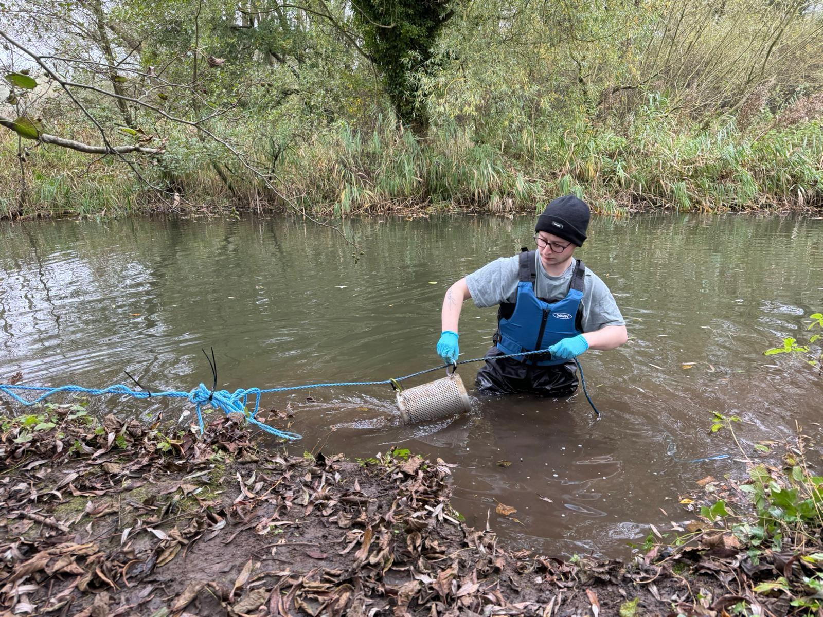 Daniel Jolly stands in a river holding small cages which contain wet wipes for his experiment.