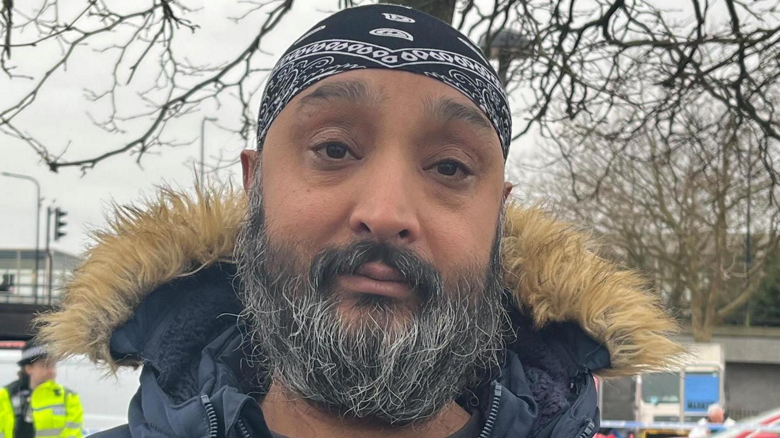 An Asian man with a beard and a black head scarf who is wearing a jumper and a coat standing in front of a police cordon with the outdoor market behind.