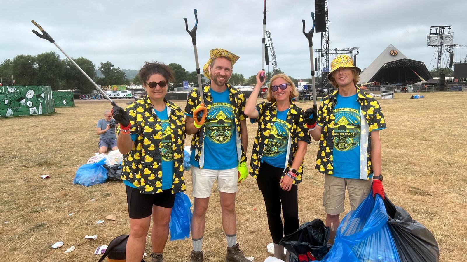 Angela Saxbee, Alastair Cullen, Sarah Parker and Gavin Eddy. Angela has dark curly hair tied up in a ponytail, Alastair has short fair hair and facial hair, Sarah has short blonde hair and Gavin has short blonde hair. Alastair and Gavin are wearing yellow hats, while Angela and Sarah are wearing sunglasses. All of them are wearing blue volunteer t-shirts and black shirts with yellow ducks on them. They are all holding litter picking sticks and bags of rubbish.