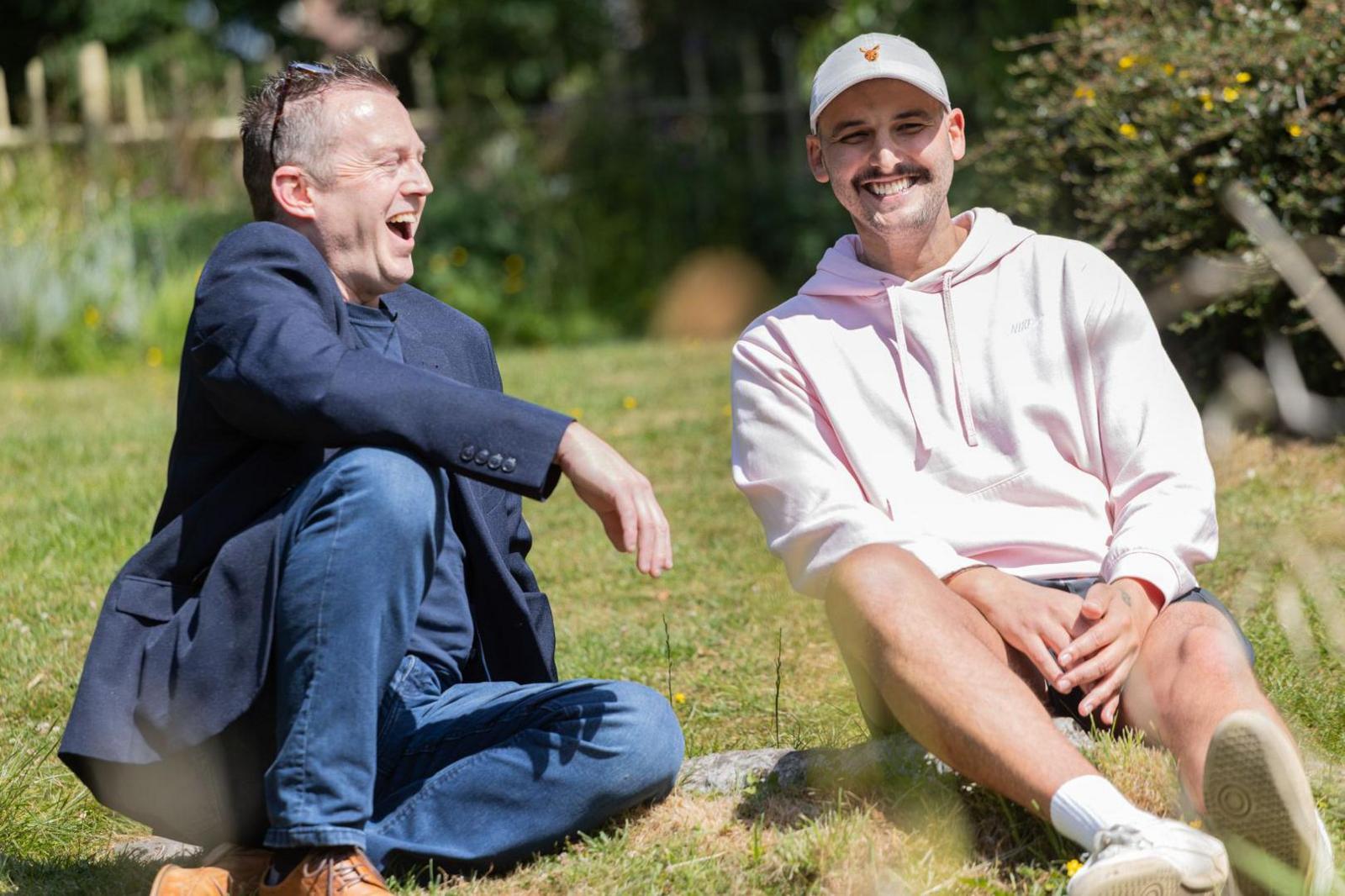 Two men sitting on grass in a park. On the left, a 51-year-old man wearing a blue blazer and blue jeans smiles broadly as he looks at a 31-year-old man who is smiling and wearing a white cap, a light-pink hoodie and shorts.