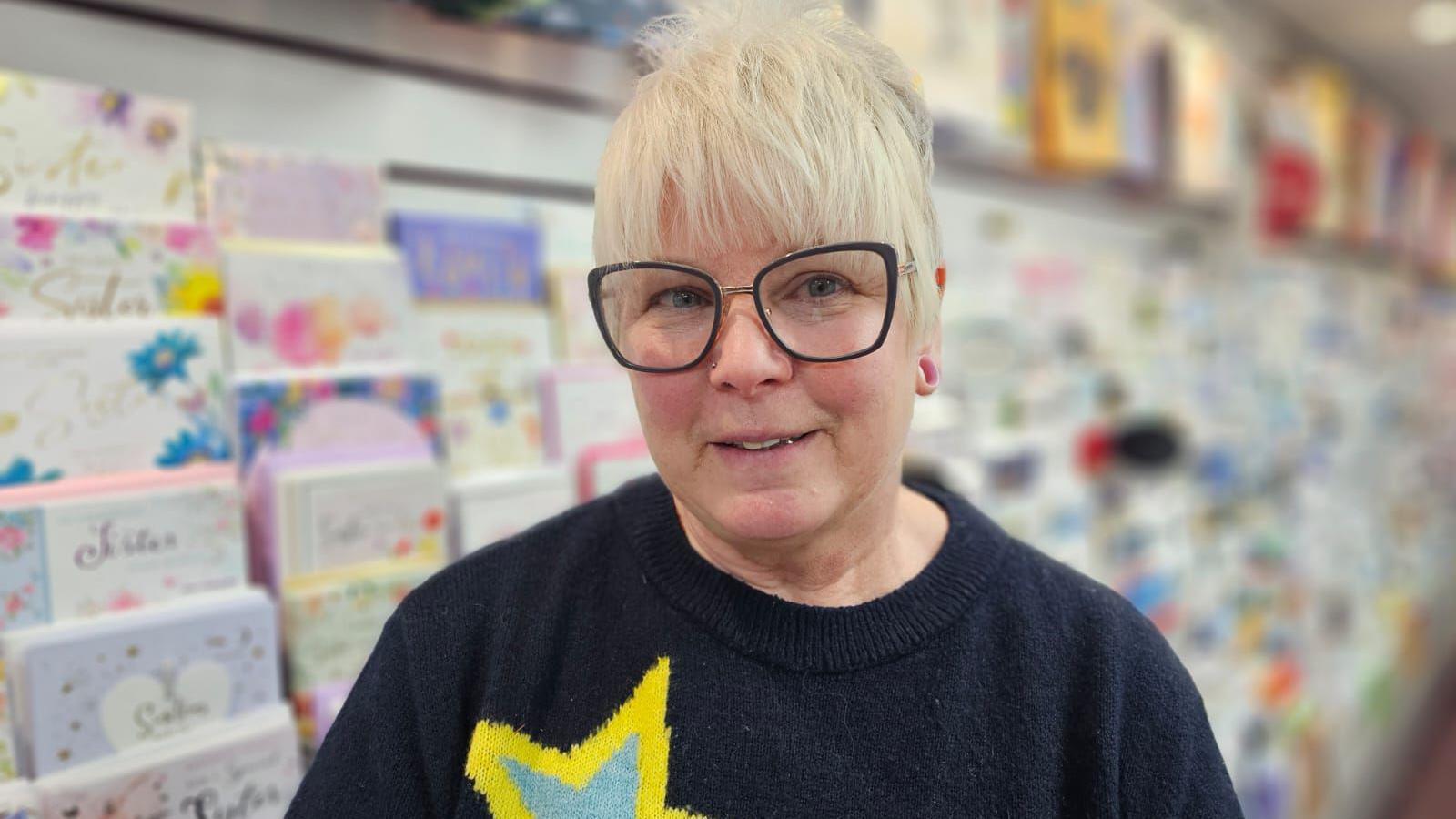 Michelle is wearing black thin framed square glasses, she has short white hair and is wearing a navy jumper with a yellow print on. She stands in front of a shelf of cards. 