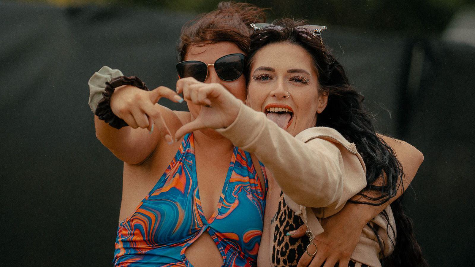 Two women pose for the camera holding one hand out each in a heart shape. They both appear to be wearing bikinis or swimming costumes.