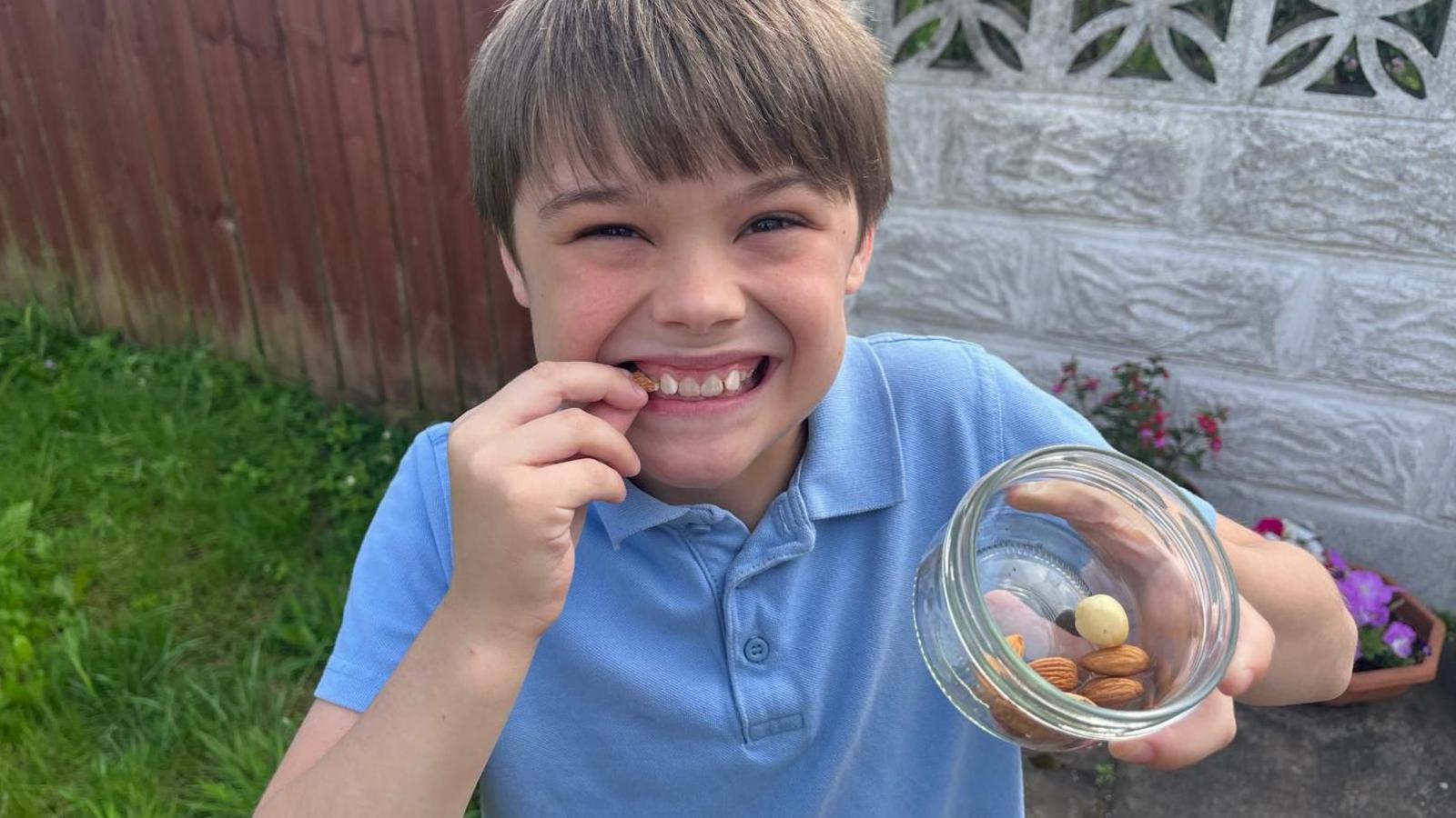A young boy wearing a blue polo-shirt, smiling with a bowl of nuts in his left hand. He's also holding a nut up to his face.  