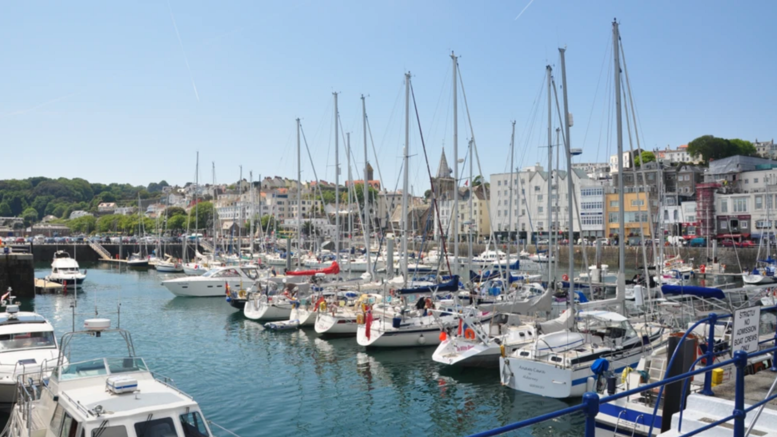 A marina with yachts and motor boards berthed with a harbour wall and buildings behind.