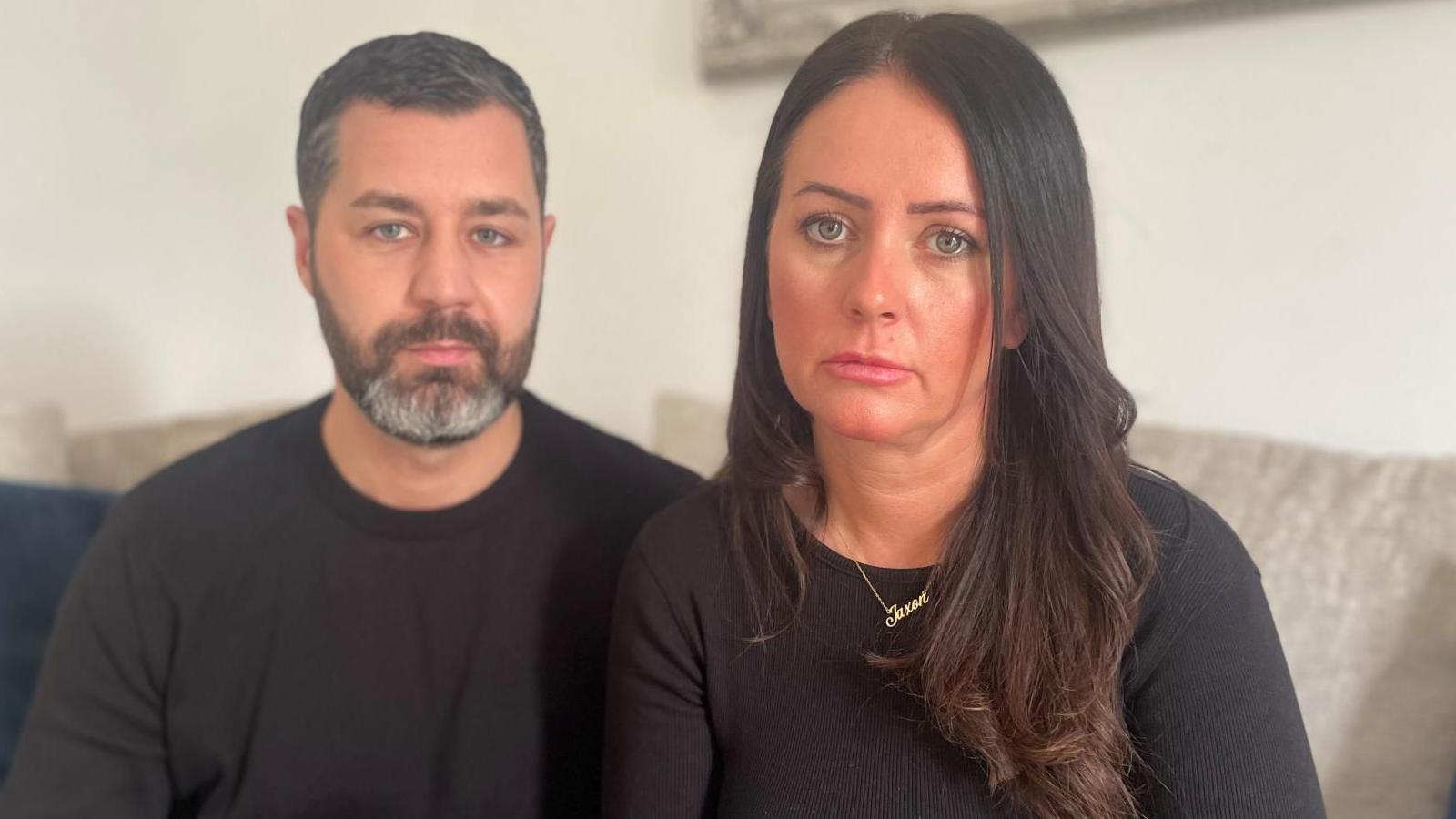 A couple dressed in black hold each other as they look towards the camera,