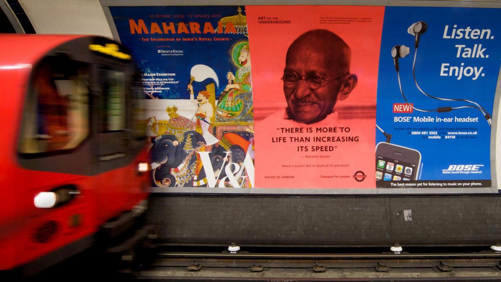 A Tube train pulling into a platform is slightly blurred at the left of the image, while on the right is a poster on the wall of the platform that has a photo of Mahatma Gandhi. There is a Gandhi quote on the poster "there is more to life than increasing its speed". 