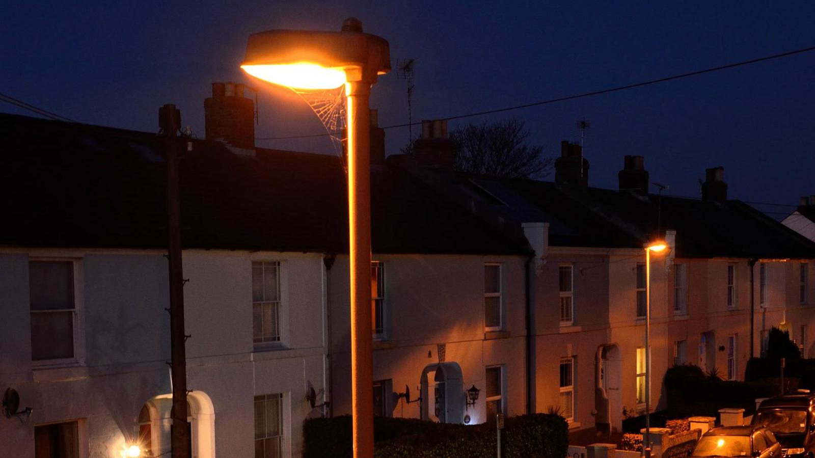 Terraced housing at night with cars parked in front. In the foreground is a street light which is on and has a large cobweb extending from it. A second street light can be seen across the road.