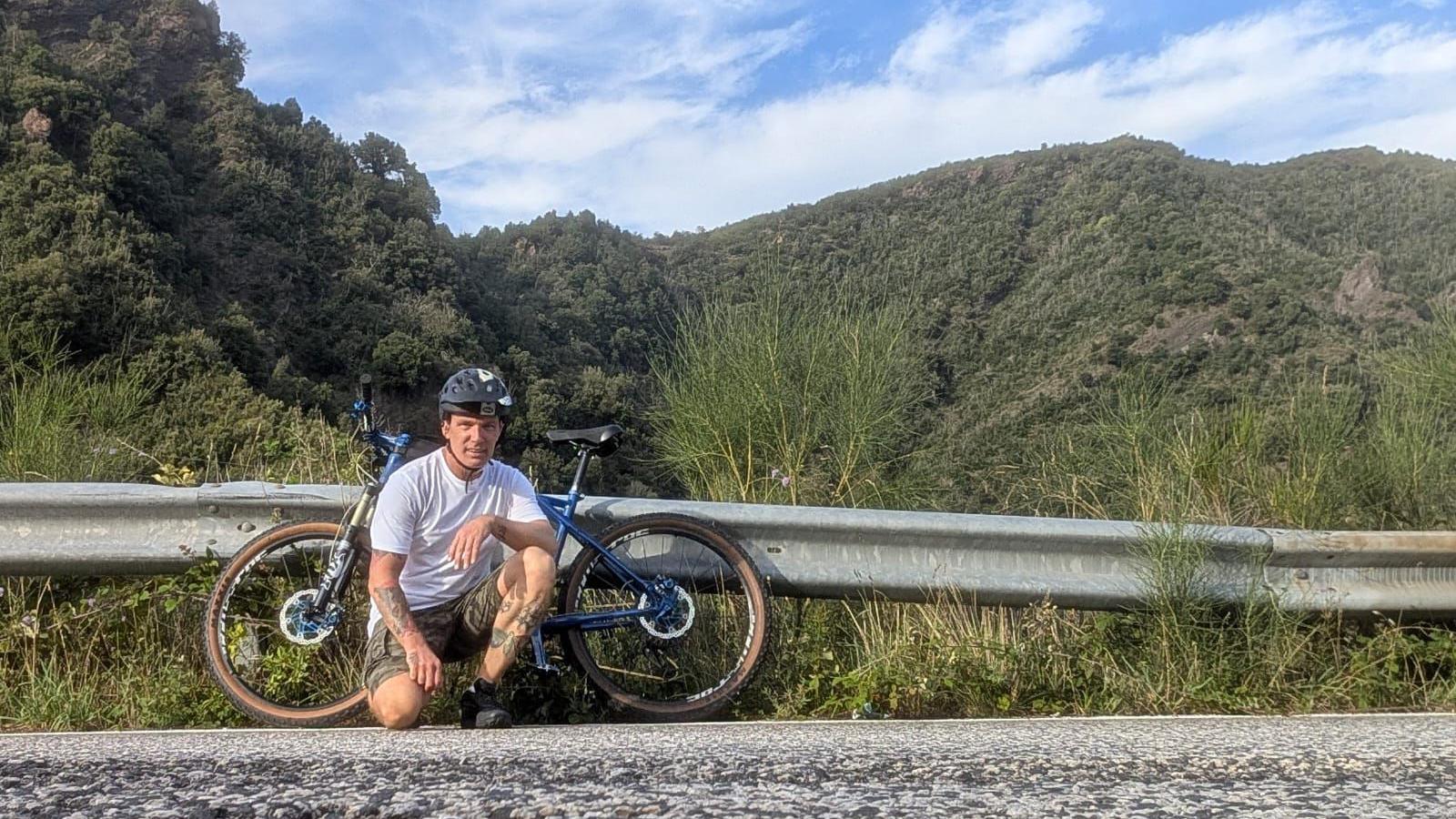 A man in a black helmet, white t-shirt, camouflage shorts and black trainers is crouching next to a blue bicycle. The bike is leaning on a metal road barrier alongside a street. Behind that is a huge mountain landscape covered in trees.