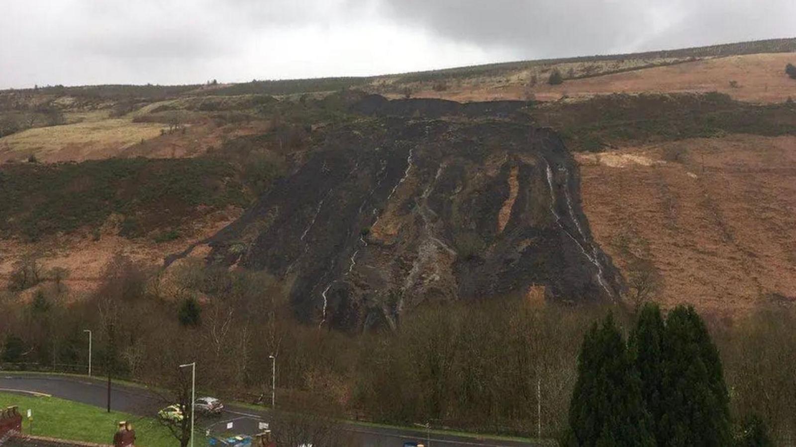 A coal tip has slipped leaving a blackened hillside. Either side of the landslip the grass and bracken is orange and yellow in colour. There is a grey, cloudy sky and in the foreground there is a road with lamp posts and trees either side of it and a couple of chimney stacks are visible in the bottom left hand corner.