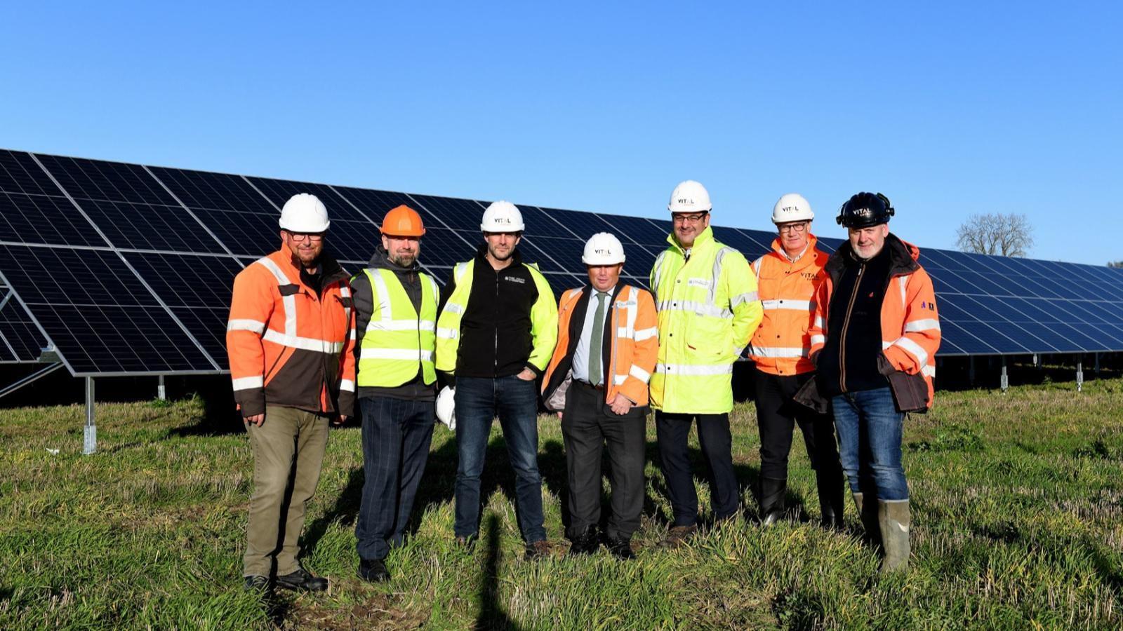 Seven men stood in a field in front of large solar panels. They are all wearing jeans, yellow or orange hi-visibility vests or jackets, and construction hats. The sky is blue.