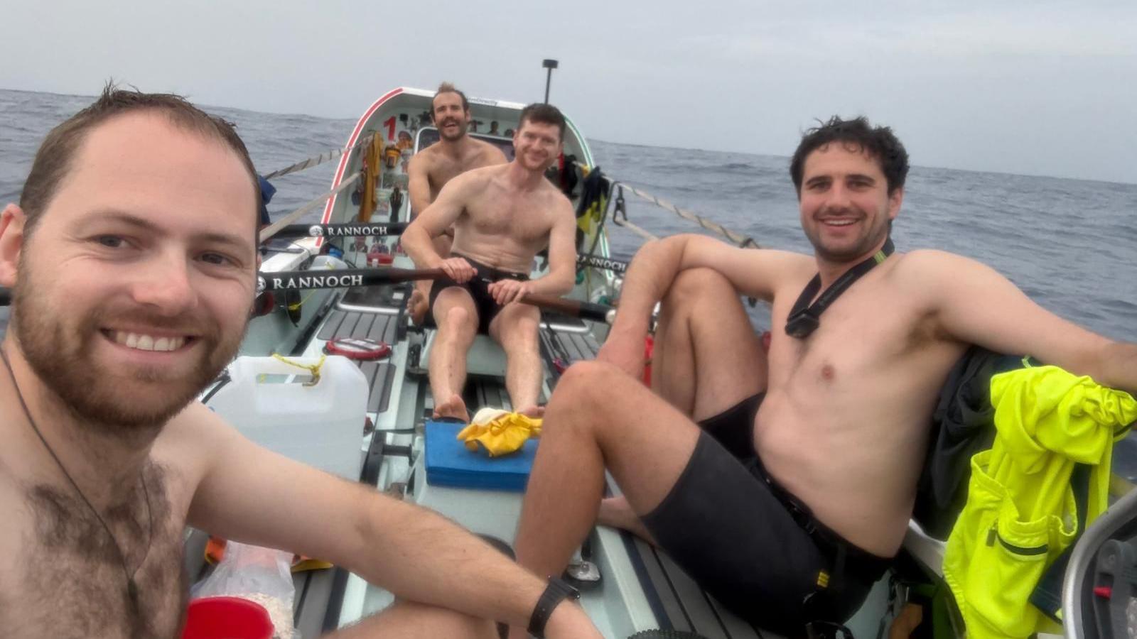 Four topless men in a rowing boat smile at the camera with the sea behind.