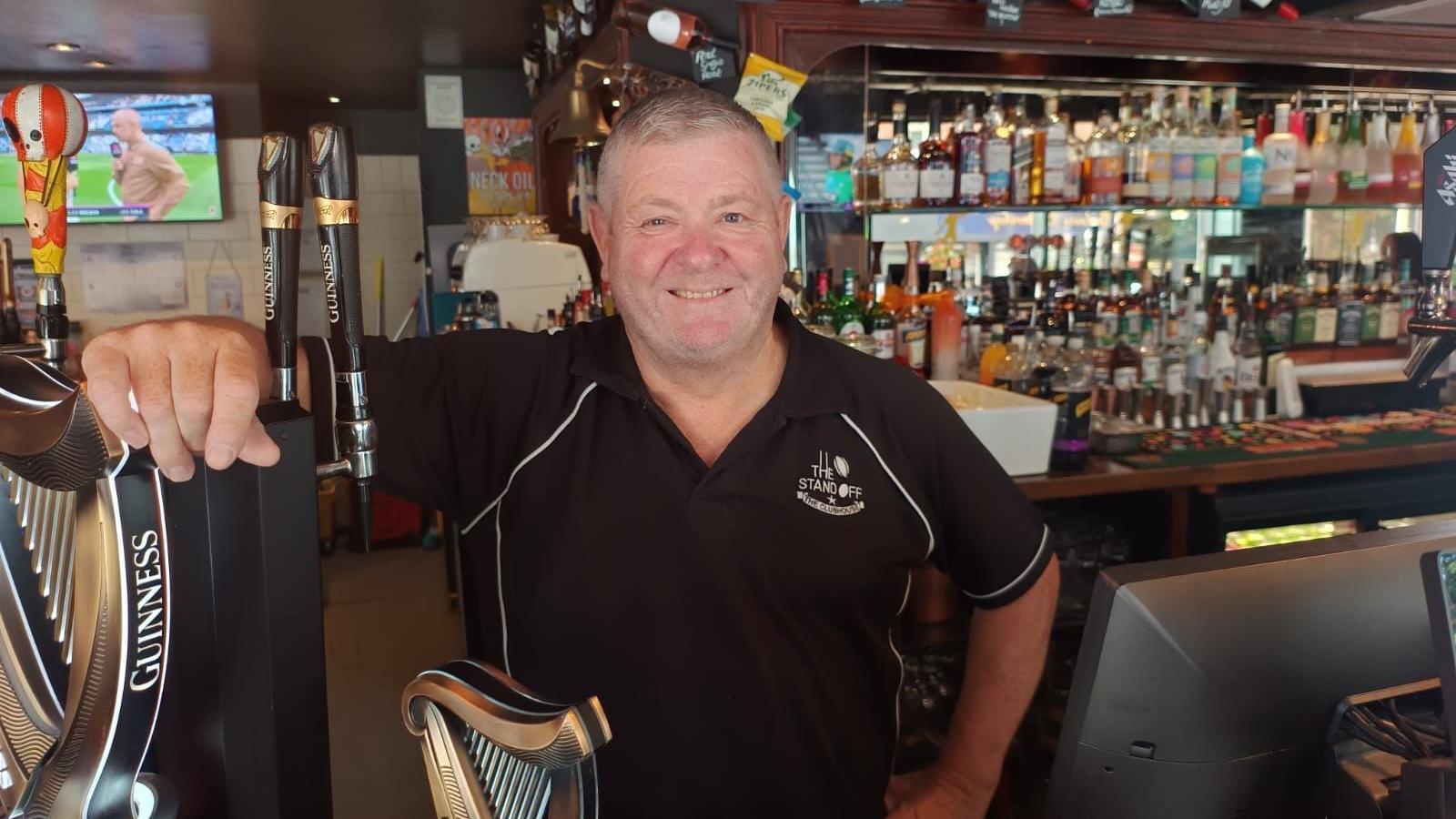 Man standing behind a bar. He is wearing a black polo top with The Standoff logo on the right. Glass alcohol bottles are lined up on the shelf behind.
