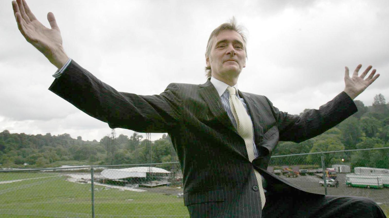 A man wearing a black pinstriped suit, yellow tie and blue and shite striped shirt is sitting with his arms outstretched. Behind him, down a hill, a stage is in the beginning stages of construction in front of a large grassy area, with trees behind it. It is an overcast day.