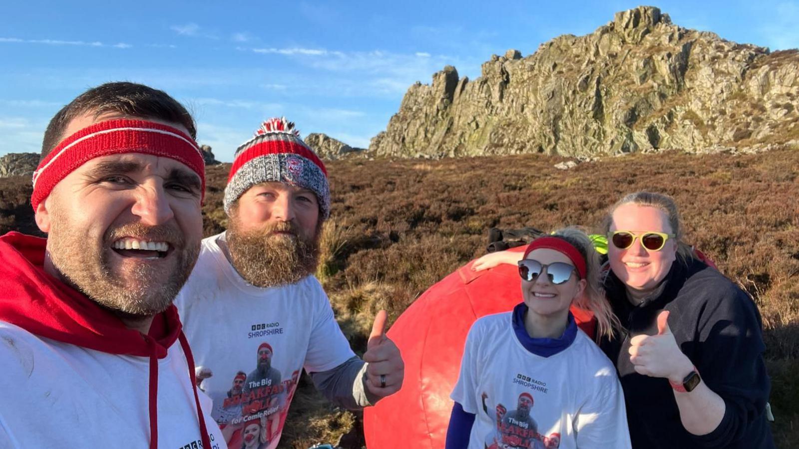 Two men and two women, wearing branded "Big Breakfast Roll" T-shirts, stand in front of the Devil's Chair rocks on Stiperstones. They are surrounded by heather and scrub under a blue sky. There is a large "red nose" made of bouncy castle fabric, which the group have pushed up the hill.