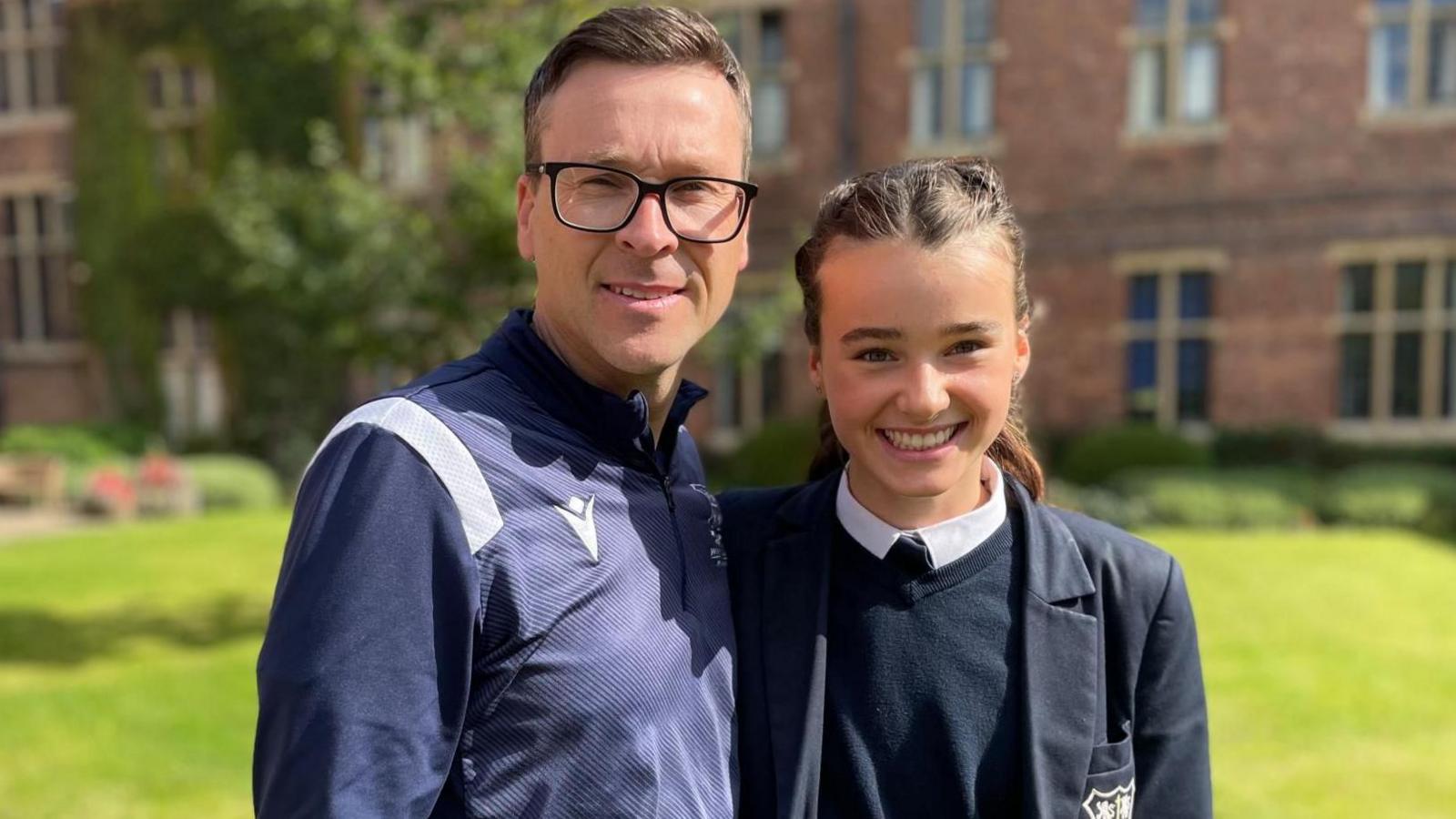 A young girl and he dad stand arm in arm smiling at the camera