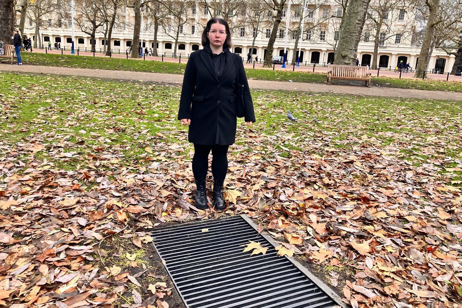 Snezana Grigorjeva is seen in a black coat stands on autumn leaves near a ground-level light grille in a city park. Behind her are leafless trees, a historic building, and a bench.