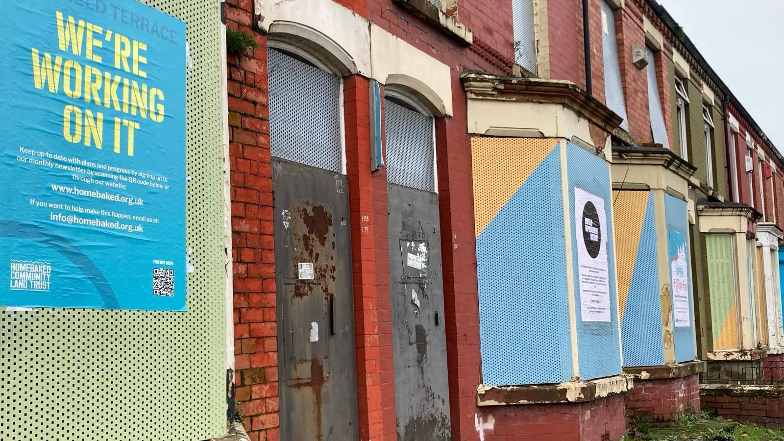 A row of terraced houses with their windows and doors shuttered with metal sheets. On one window is a poster saying 'We're working on it' with website details for Homebaked CLT,