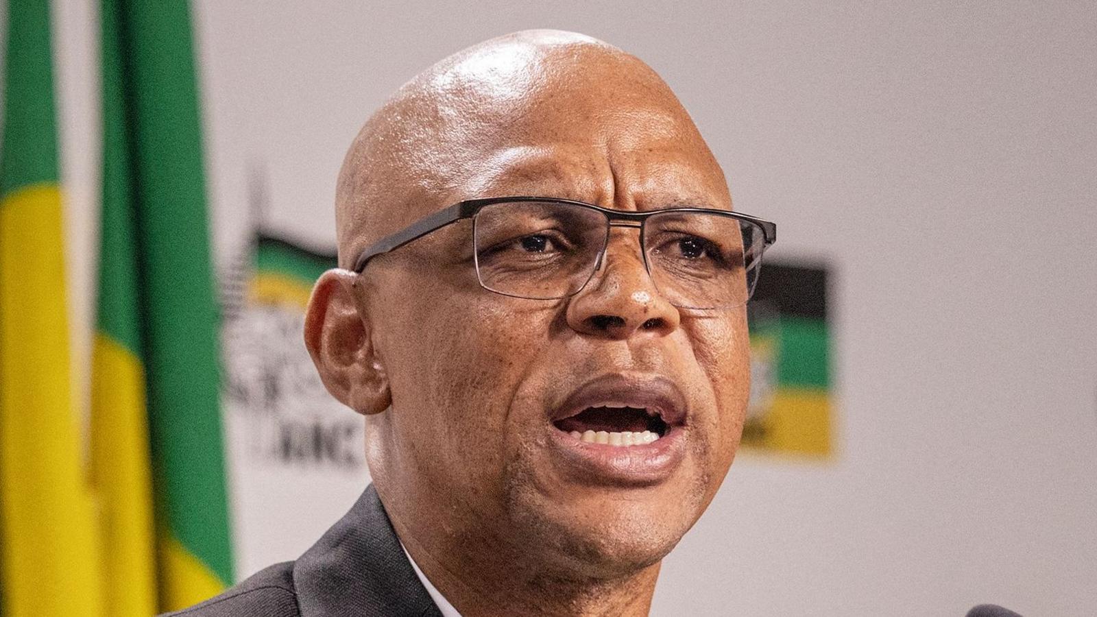 Pule Mabe - wearing a pair of glasses - addresses a press conference. ANC colours can be seen in the background.