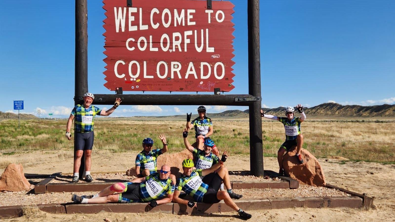 The group of cyclists in front of a sign saying 'welcome to colourful Colorado' surrounded by sand and grass