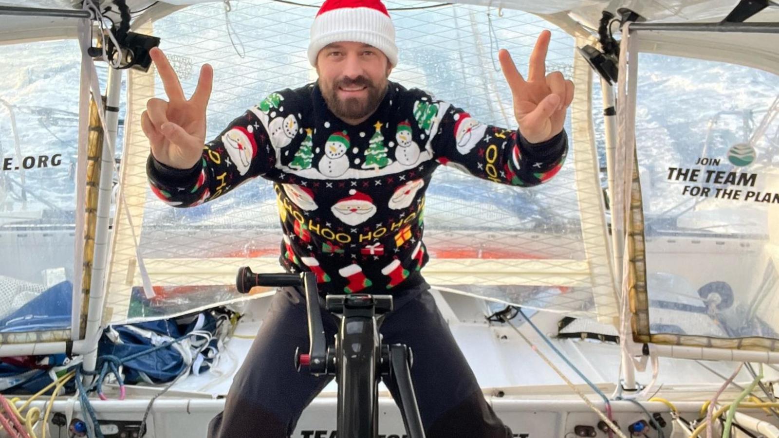 A man in a Christmas jumper and hat gives two V for victory signs in the cockpit of a yacht.