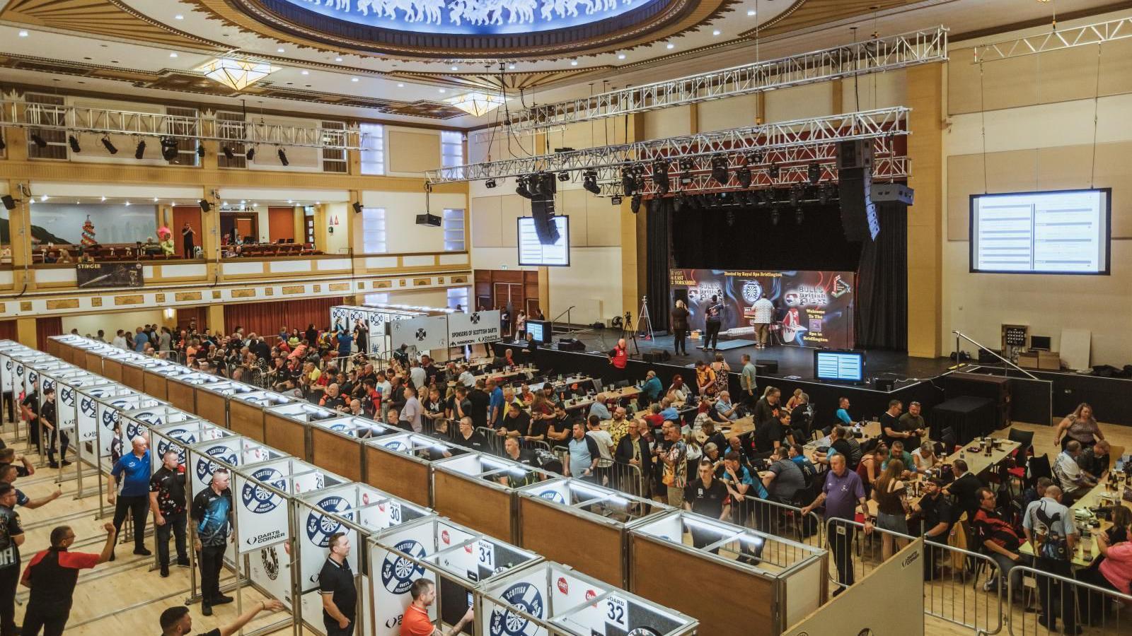 Interior of Bridlington Spa with rows of partitioned dart boards in the foreground in front of rows of desks with dozens of people sitting at them and a raised stage in the background