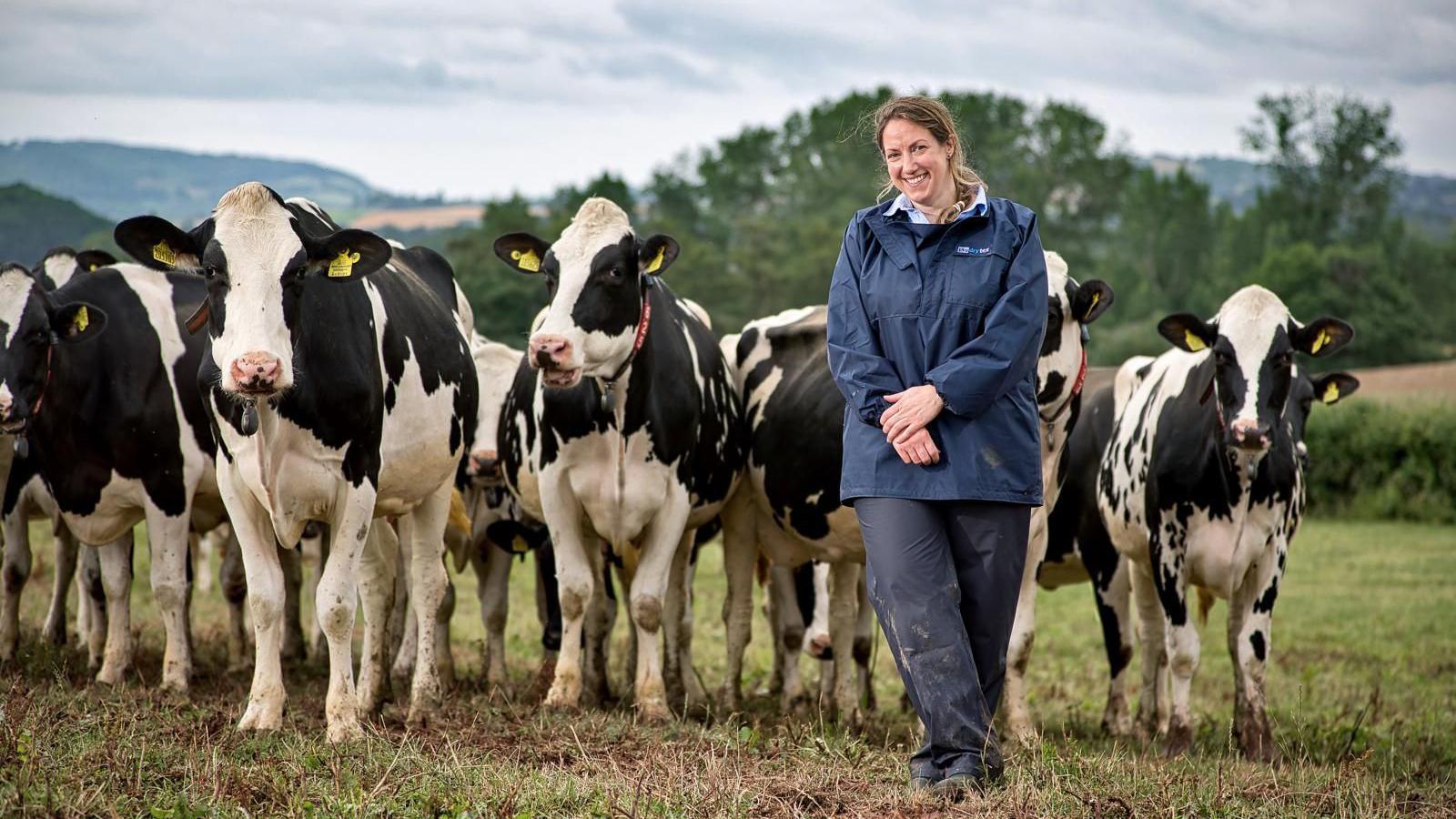 Farm vet Sara Pedersen stands looking direct to camera in front of some cows. She is wearing a blue coat and waterproof trousers.
