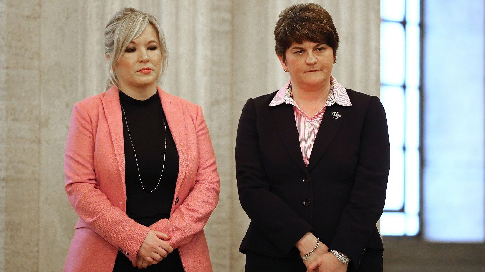 Michelle O'Neill standing next to Arlene Foster. O'Neill has blonde hair tied back with a fringe wearing a pink blazer and black top, and a long silver necklace. She is wearing makeup with pink lipstick. Foster has short brown hair and is wearing a pink striped shirt with a black blazer.