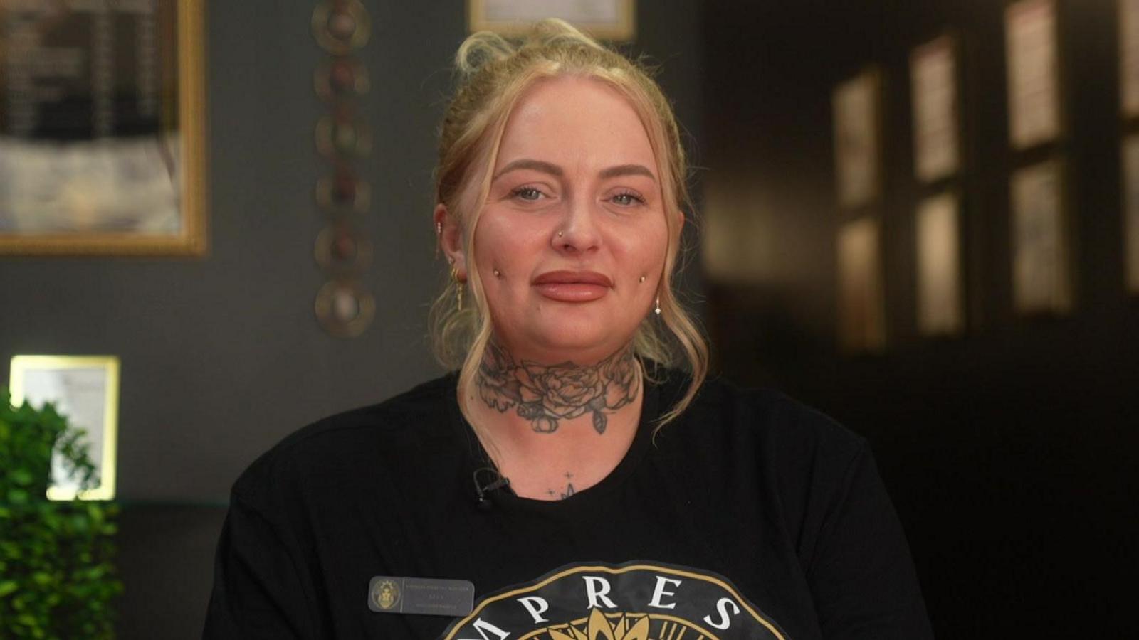 Lucy is sat in a professional piercing studio. Her blonde hair is tied up with two pieces loose, framing her face. She has a floral neck tattoo and piercings on each side of her cheeks and nose. She also has several ear piercings. She is wearing a black t-shirt with a logo on the front. An employee badge is place on the left side of her t-shirt.