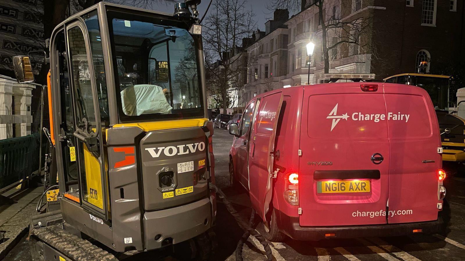 A Volvo branded, grey and yellow construction vehicle being charged by a red van with the branding 'Charge Fairy' on a road. 