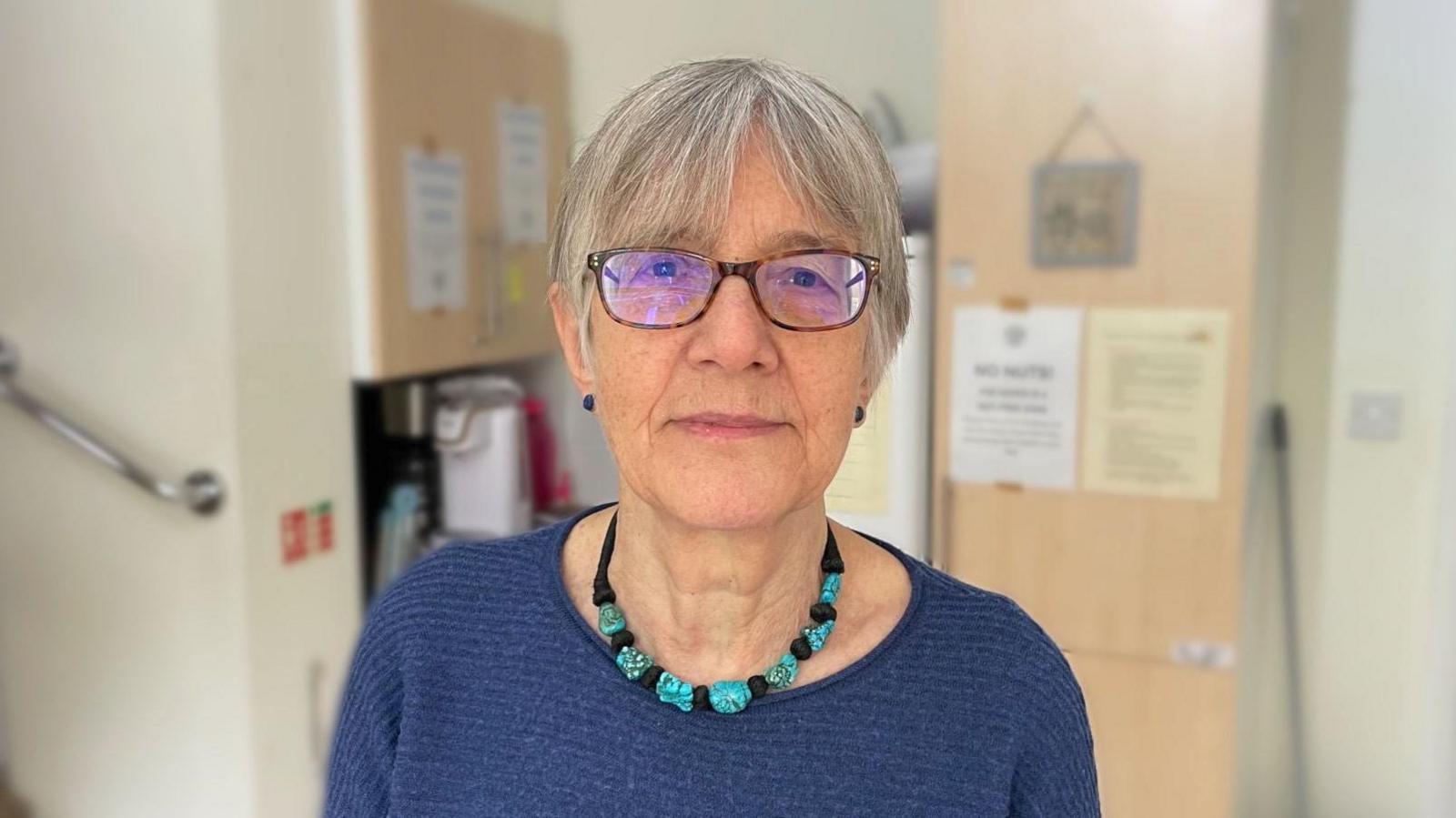 Diana Minns, wearing glasses, a blue knitted top, and a black and turquoise necklace, is looking directly at the camera. She is standing in front of cupboards and a fridge in the kitchen.