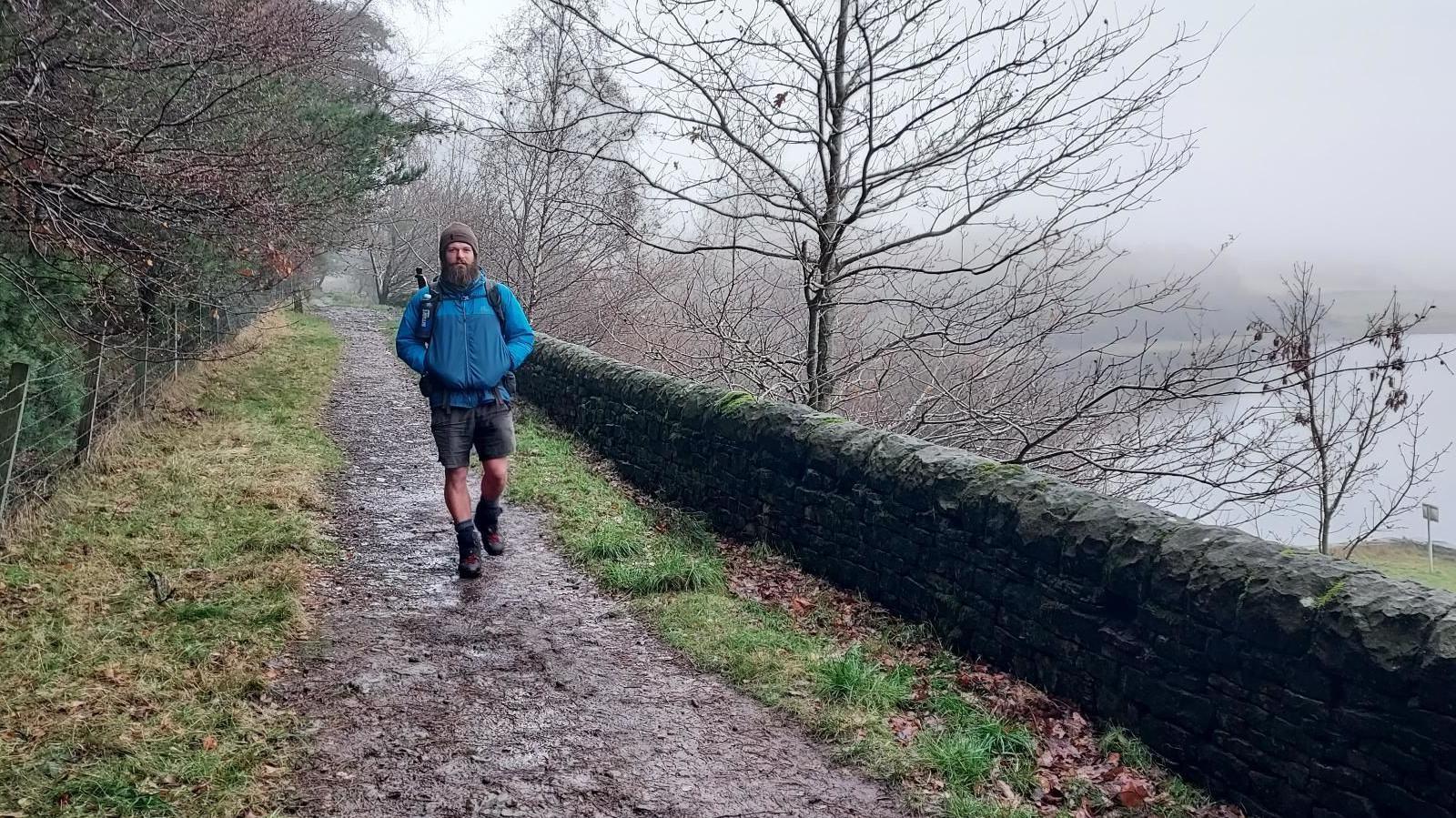 A man in shorts, brown boots and a blue coat walks along a muddy track along a stone wall. Trees can be seen to one side of the track, while on the other, a slope runs down to the water of a reservoir. 