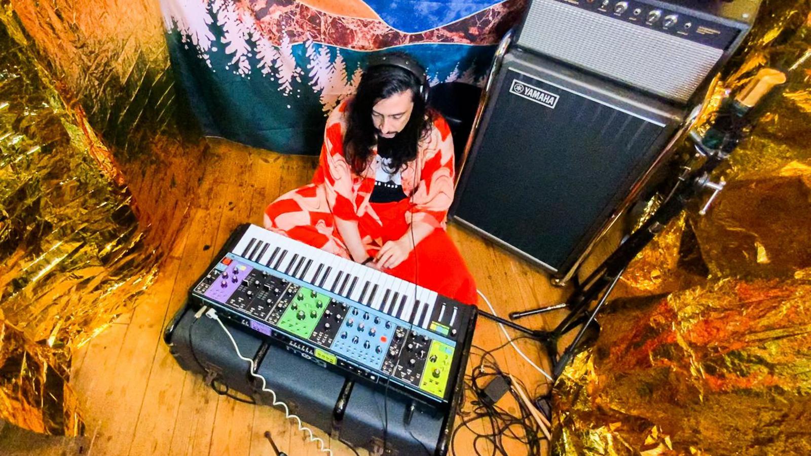 A shot of Will from above in a small recording space, which has gold foil curtains hung on both sides and a batik style drape of mountains and forests behind him. He sits cross legged on a wooden floor, in front of a large electric keyboard and synth. He wears headphones and sits next to a large Yamaha speaker and amp. He wears bright red flowing jacket, bright red trousers and a black T-shirt.