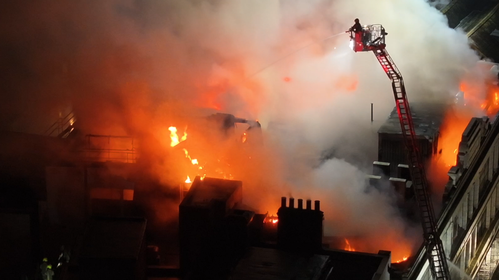 A drone shot of someone on an aerial platform shooting water at flames on the roof of the building.