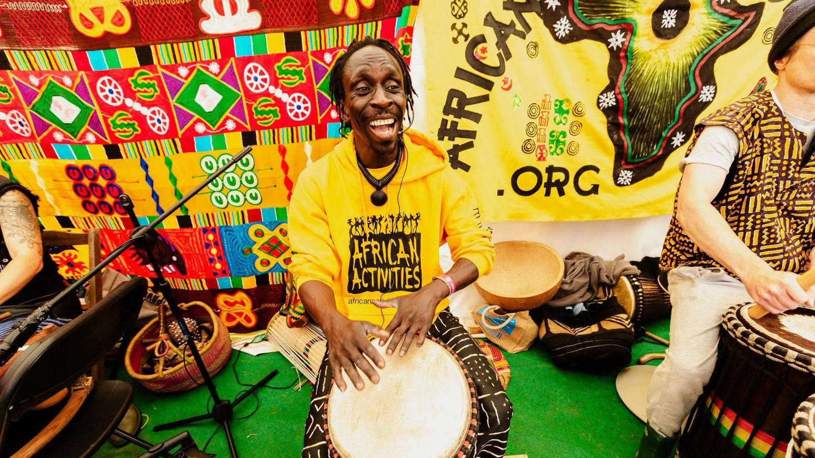 A black man plays a drum between his legs with his hands. He wears a yellow T-shirt with black writing saying "African Activities".