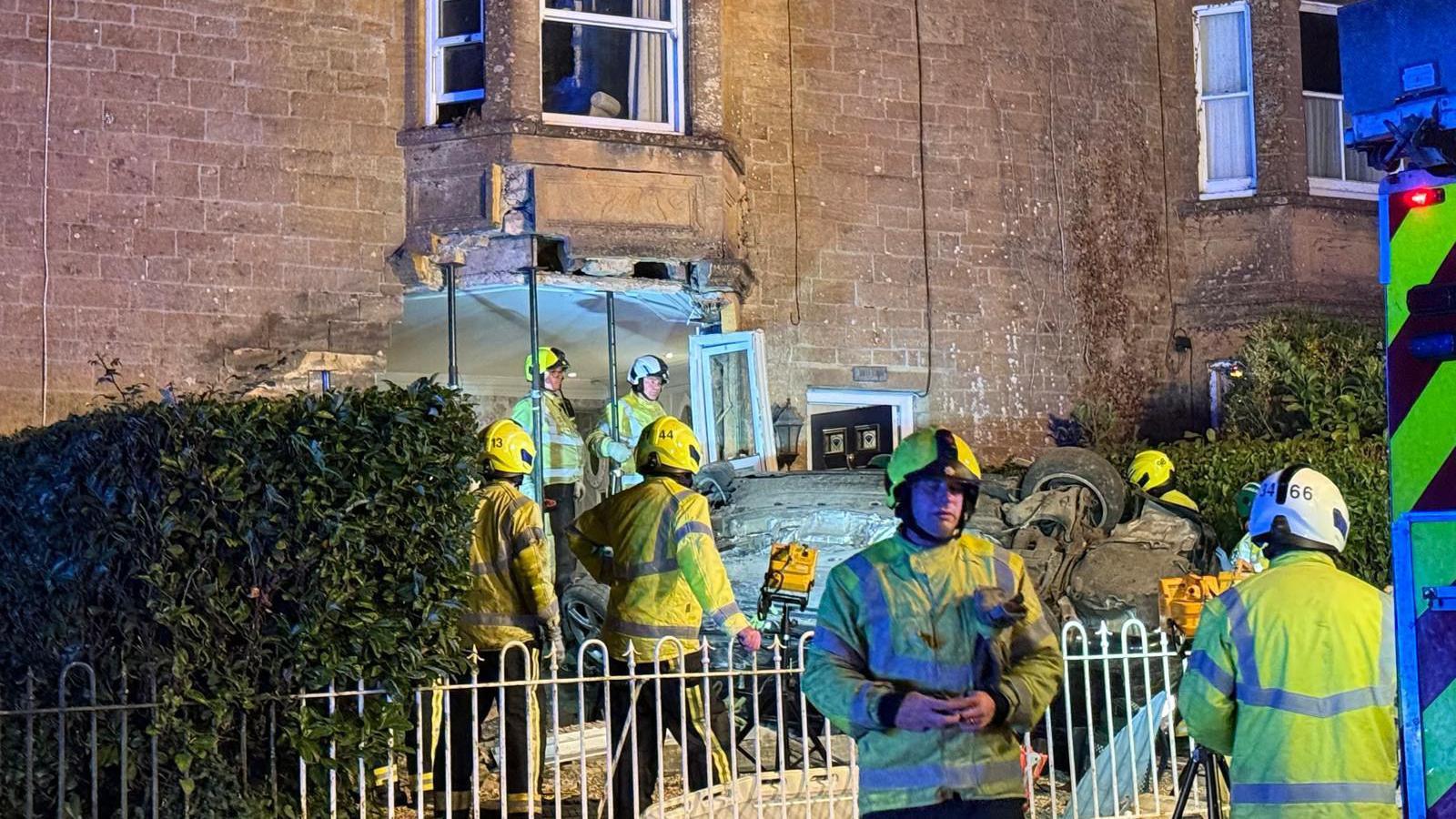 A number of fire crew personnel stand in front of the house at the scene of the incident. Most of them are looking at the car, which is on its back. The house is lit with industrial lighting.